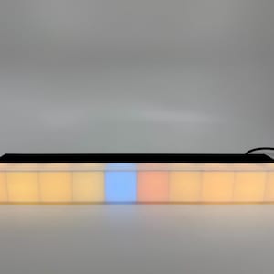 May include: A rectangular LED light bar with a black top and a series of illuminated, square-shaped panels. The panels display a gradient of colours, including yellow, blue, and orange. A black cable extends from the right side.
