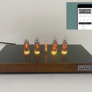 May include: A wooden clock with a black face and five orange glowing Nixie tubes displaying the time 11:20. The clock has a small white button on the left side and a silver ATHNH logo on the right side. The clock is connected to a black power cord.