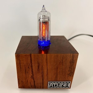 May include: A small, clear glass tube with a glowing orange light inside, sitting on a wooden base with a blue light at the bottom. The base is made of dark wood with a grain pattern and has a metal label with the text "ATHNK" on the front.