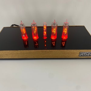 May include: A red digital clock with six Nixie tubes displaying the time 11:44. The clock is on a black and wood base with the word "ATHN" on the front.