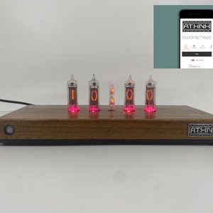 May include: A wooden clock with five red glowing Nixie tubes displaying the time "10:00". The clock has a black base and a silver label with the text "ATHNK" on the front. The clock is connected to a power source with a black cable.