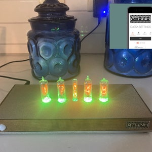 May include: A wooden clock with six green and orange Nixie tubes displaying the time 17:48. The clock is on a table with two blue glass jars in the background. A smartphone with the text "ATHNH CLOCK SETTINGS" is visible in the top right corner of the image.