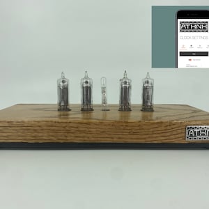 May include: A wooden clock with four clear glass Nixie tubes displaying the time. The clock has a black base and a silver metal logo that says "ATHNK".
