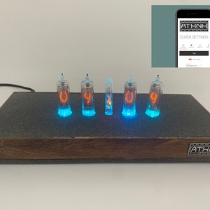 May include: A digital clock with five glowing glass tubes displaying numbers in orange. The clock sits on a dark gray surface with a wooden frame. The clock's brand name, ATHNH, is visible. A smartphone screen shows clock settings.