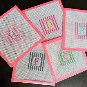 May include: Five square needlepoint canvases with pink fabric borders. Each canvas features a central letter monogram surrounded by vertical stripes. The letters include H, D, B, F, and C, rendered in various colors including pink, orange, teal, and green.