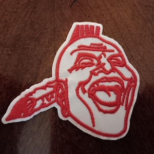 May include: Embroidered patch featuring a stylized Native American head in red on a white background. The design includes a feathered headdress and an open mouth, with a red outline.