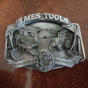 May include: Silver belt buckle with a 3D design of a farm scene, including a tractor, a barn, and a person with a wheelbarrow. The buckle has the text "AMES TOOLS" and "HELPING AMERICA GROW SINCE 1774" on it.