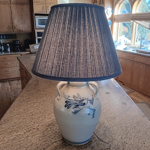 May include: A ceramic lamp with a blue pleated lampshade. The white ceramic base features blue bird and floral designs. The lamp has two handles and sits on a granite countertop. The lamp is plugged into an electrical outlet.