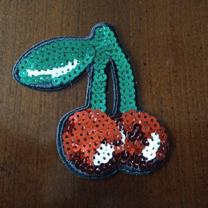 May include: A red and green sequined cherry patch. The cherry is made of red and white sequins, and the stem and leaf are made of green sequins.