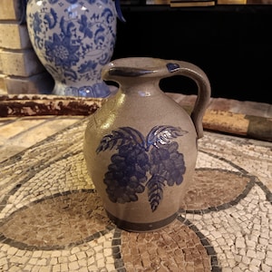 May include: A small, gray stoneware pitcher with a blue and white grape design. The pitcher has a handle and a rounded body.
