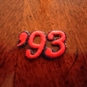 May include: A fabric patch featuring the number 93 embroidered in bright red thread with a dark outline. The numerals are connected and have a raised, textured appearance, creating a distinct graphic element suitable for application onto textiles.
