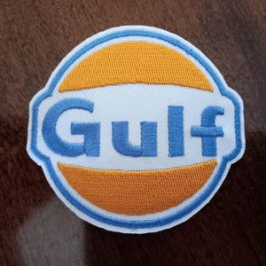May include: Embroidered patch featuring the Gulf logo. The patch has a white background with a blue border. The word "Gulf" is embroidered in blue, with orange and white accents. The patch is circular with a unique shape.