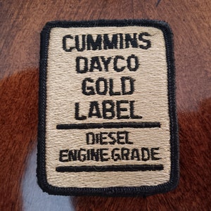 May include: A beige fabric patch with black embroidered text that reads "Cummins Dayco Gold Label Diesel Engine Grade".