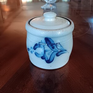 May include: A small, cylindrical, off-white stoneware jar with a matching lid. The jar features a hand-painted blue floral motif on the front. The surface has a speckled texture, and the lid includes a small, rounded knob handle at the center.