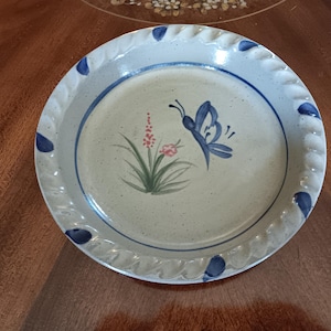 May include: A round ceramic pie dish with a speckled off-white glaze. The rim features a decorative rope-like texture with painted blue accents. The center displays a hand-painted design of a blue butterfly, green grass, and small pink flowers.