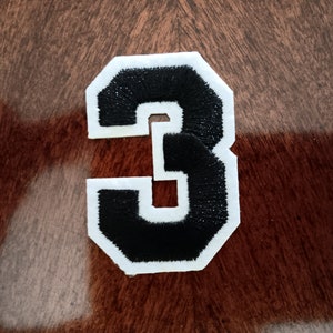 May include: A black felt number "3" with white stitching, on a brown wooden background.