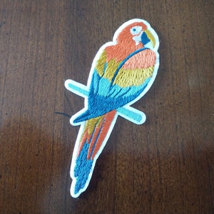 May include: Embroidered iron-on patch of a colorful parrot with blue, orange, and yellow feathers, perched on a branch.
