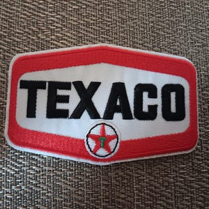 May include: A rectangular embroidered patch with rounded corners featuring a white center and a thick red border. The word TEXACO is stitched in bold black capital letters across the middle, with a small red star logo containing a green T below.