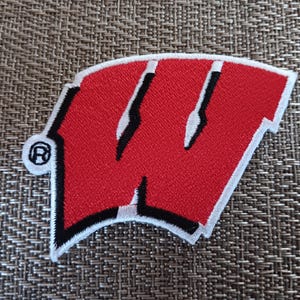 May include: Embroidered patch featuring a red "W" with black and white accents, a white border, and a registered trademark symbol. The patch is in the shape of a shield. The patch is likely a team logo.