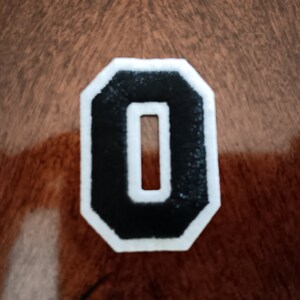 May include: A black felt number zero with white outlining, in a varsity style font. The number is on a brown wooden surface.
