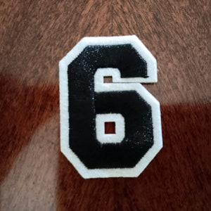 May include: A white felt patch with a black felt number six. The number is outlined in white.