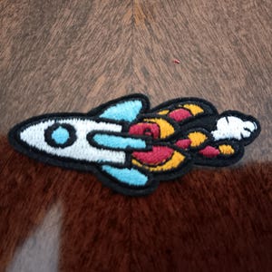 May include: Embroidered patch of a white rocket ship with blue and black accents, and colorful exhaust flames in red, yellow, and white. The patch is on a brown and tan textured surface.