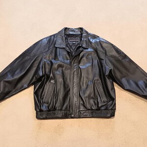 May include: A black leather jacket with a classic design. The jacket features a collared neckline, a front zipper closure, and two front pockets. The jacket has a ribbed hem and cuffs. The brand name is visible on the label.