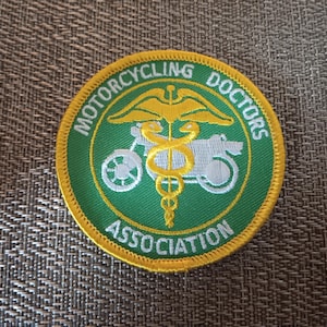 May include: A round patch with a green background and a yellow border. The patch features the words "MOTORCYCLING DOCTORS ASSOCIATION" in a circular pattern. A motorcycle and a caduceus are in the center.