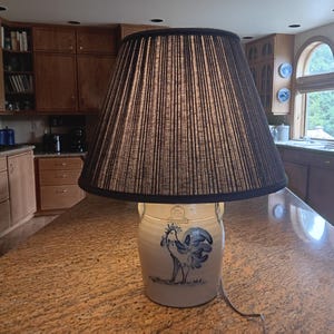 May include: A table lamp featuring a cream-colored ceramic crock base decorated with a blue hand-painted rooster illustration. The lamp is topped with a large, conical fabric lampshade in a dark charcoal gray with fine vertical pinstripes.