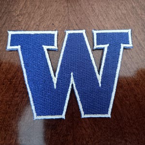 May include: A blue embroidered patch in the shape of the capital letter W. The letter features a thick, dark blue center with a white border outline. The embroidery has a visible textured stitch pattern throughout the entire surface of the letter.