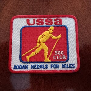 May include: A white and red embroidered patch with the text "USSA 500 CLUB" and "KODAK MEDALS FOR MILES". A yellow silhouette of a person cross-country skiing is in the center of the patch.