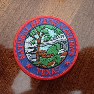 May include: A round patch with a red border and blue center featuring the text "NATURAL BRIDGE CAVERNS TEXAS". The patch has an embroidered design of a tree, cave, and landscape in green, brown, and white.