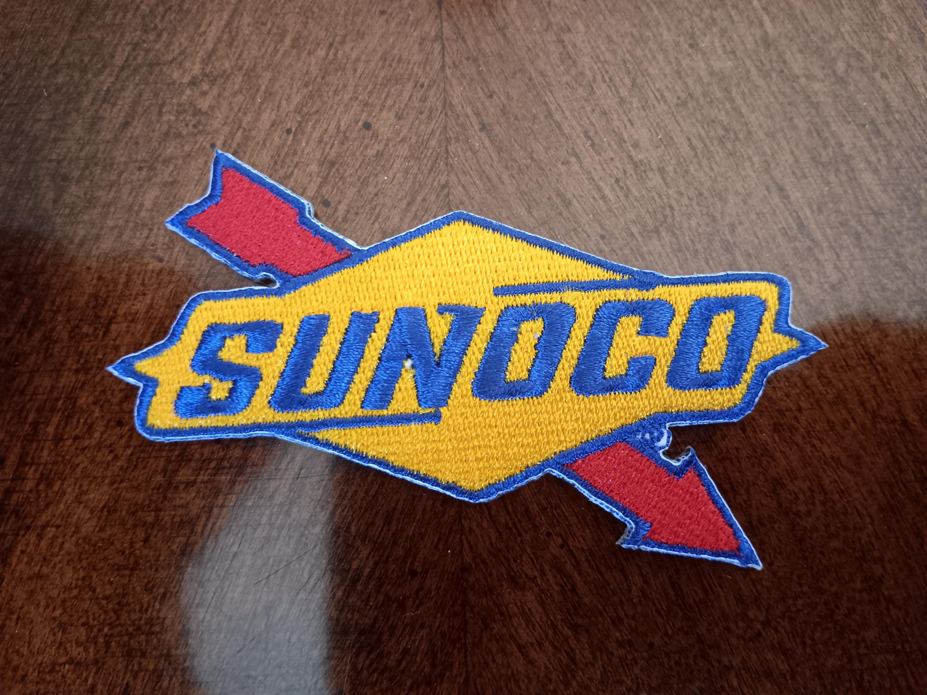 May include: An embroidered fabric patch featuring the Sunoco logo. The design consists of a yellow diamond shape with a blue border, containing the blue text SUNOCO. A red arrow with a blue outline extends diagonally behind the diamond.