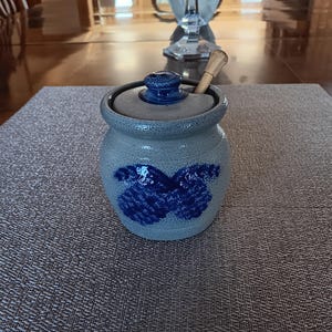 May include: A blue and gray ceramic honey pot with a butterfly design. The pot has a lid and a wooden honey dipper.