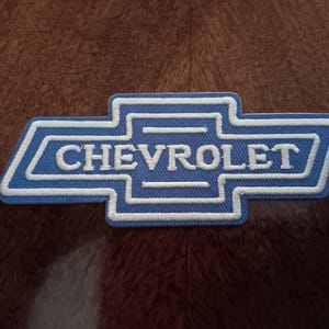 May include: A blue and white embroidered patch featuring the Chevrolet logo. The design consists of the iconic bowtie shape outlined in white thread, with the word CHEVROLET stitched in white capital letters across the center of the blue background.