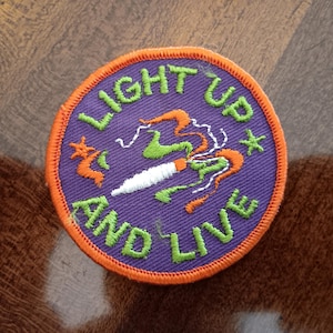 May include: Embroidered patch with the words "LIGHT UP AND LIVE" in green lettering on a purple background. The patch features a white object with green and orange smoke-like designs, and an orange border.