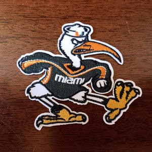 May include: An embroidered patch featuring a white ibis mascot wearing a black jersey with orange trim and the word Miami in white text. The ibis has a long orange beak, a white sailor hat with an orange logo, and yellow feet with black claws.