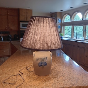 Puede incluir: Lámpara de mesa decorativa con una pantalla plisada marrón y una base de cerámica. La base es blanquecina con diseños florales azules y dos pequeñas asas. La lámpara está sobre una encimera de granito.