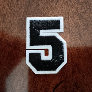May include: Black embroidered number 5 patch with a white border. The number is in a bold, block style with a stitched texture. The patch is likely for a sports uniform or apparel.