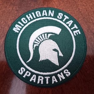 May include: A circular patch with a green background and white embroidered details. The text "MICHIGAN STATE" arches across the top, and "SPARTANS" is at the bottom. A white Spartan helmet is in the center.