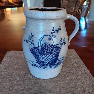 May include: A beige stoneware crock with a single handle and a wide rim. The front features a blue painted design of a woven basket filled with berries, with additional berry sprigs extending upward and scattered around the base of the basket.