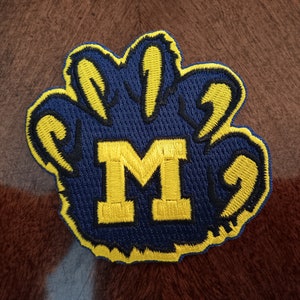May include: An embroidered patch in the shape of a paw print with dark blue thread and a bright yellow border. The center of the paw print features a large, bold yellow letter M stitched in a block font, contrasting against the dark blue background.