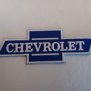 May include: A rectangular embroidered patch featuring the blue Chevrolet bowtie logo with white stitched borders and the word CHEVROLET in white capital letters centered across the middle of the emblem on a textured white surface.
