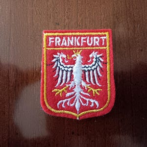 May include: Embroidered patch featuring the word "FRANKFURT" above a stylized white eagle with black and white wings on a red shield. The patch has a yellow border and is likely a souvenir or emblem.