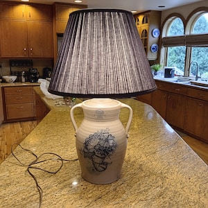 Puede incluir: Una lámpara de mesa decorativa con una pantalla plisada marrón oscuro y una base de cerámica color crema. La base presenta un diseño floral azul y dos asas. La lámpara está colocada sobre una encimera de granito en una cocina.