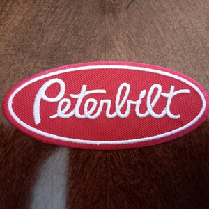 May include: An oval-shaped embroidered patch featuring a red background with a white border and the word Peterbilt stitched in white script lettering across the center. The patch has a textured fabric finish and a distinct diagonal weave pattern.