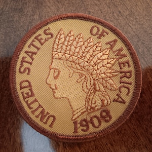 May include: A circular patch with a tan background and a brown border. The patch features a profile of a Native American in a headdress, with the text "UNITED STATES OF AMERICA 1909" in brown lettering.