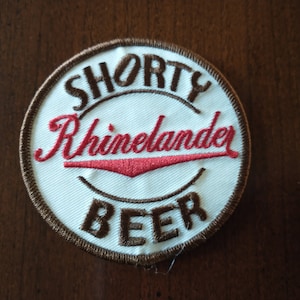 May include: Circular patch with the words "SHORTY Rhineland BEER" embroidered in brown and red on a white background. The patch has a brown border and is set against a dark wood grain surface.