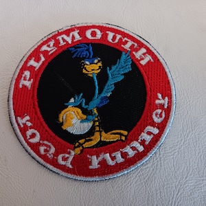May include: A circular embroidered patch featuring a black center with a blue and yellow Road Runner cartoon character. The patch has a wide red border with the white embroidered text PLYMOUTH ROAD RUNNER encircling the central graphic.