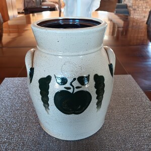 May include: A white ceramic crock with green and brown painted apple design. The crock has two handles and a brown rim.